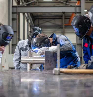 5 welders in a shop working, one with a flexed back and neck